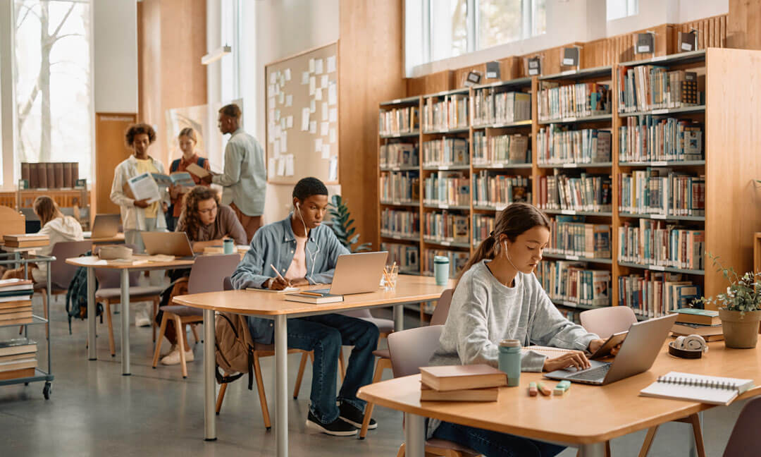 Students study, read, and use laptops in a bright school library with bookshelves and collaborative work areas.